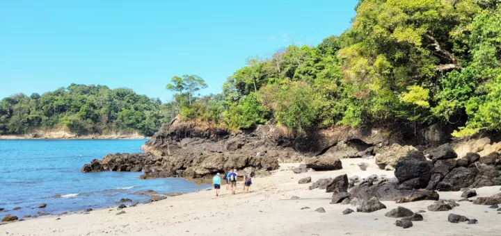 Tres personas caminan por una playa de arena clara entre rocas oscuras y selva verde, con el mar azul a la izquierda