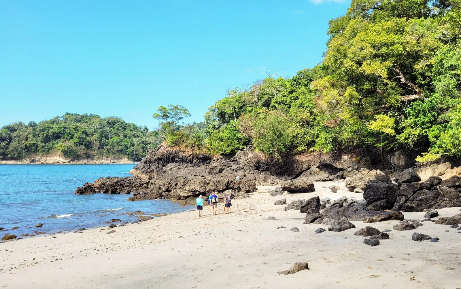 Tres personas caminan por una playa de arena clara entre rocas oscuras y selva verde, con el mar azul a la izquierda