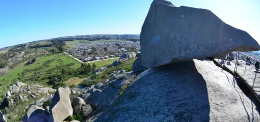 Gran roca apoyada sobre un cerro de Tandil, con campos verdes, la ciudad y un cielo azul de fondo