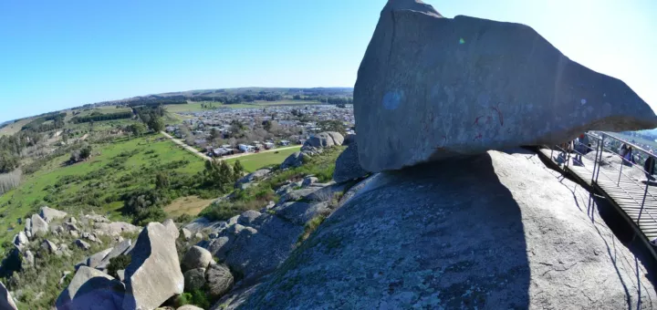 Gran roca apoyada sobre un cerro de Tandil, con campos verdes, la ciudad y un cielo azul de fondo