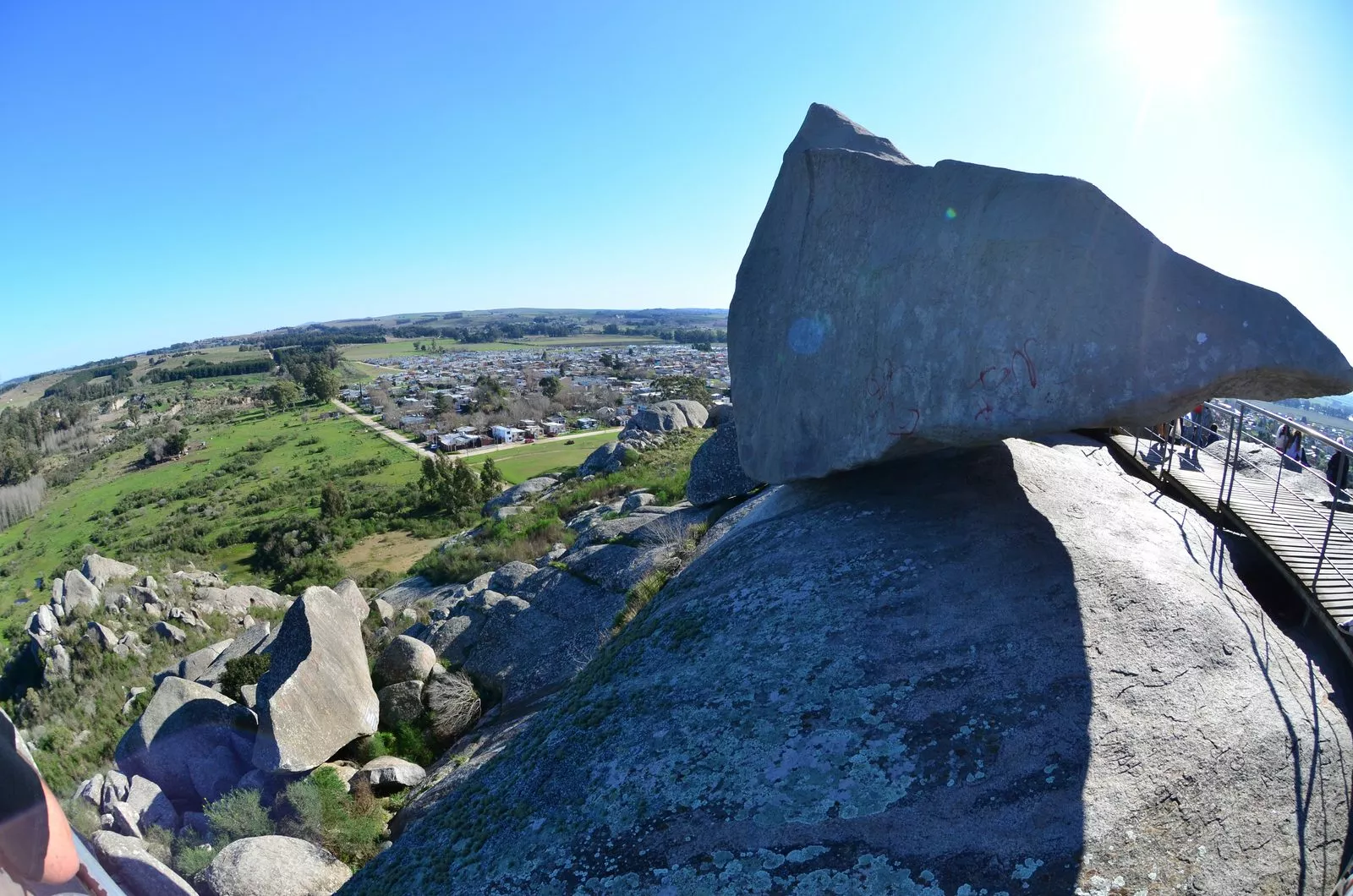Gran roca apoyada sobre un cerro de Tandil, con campos verdes, la ciudad y un cielo azul de fondo