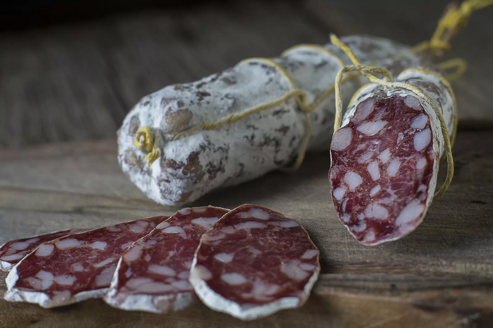 Salame curado atado con hilo amarillo, una pieza cortada al medio y varias rodajas sobre una tabla de madera