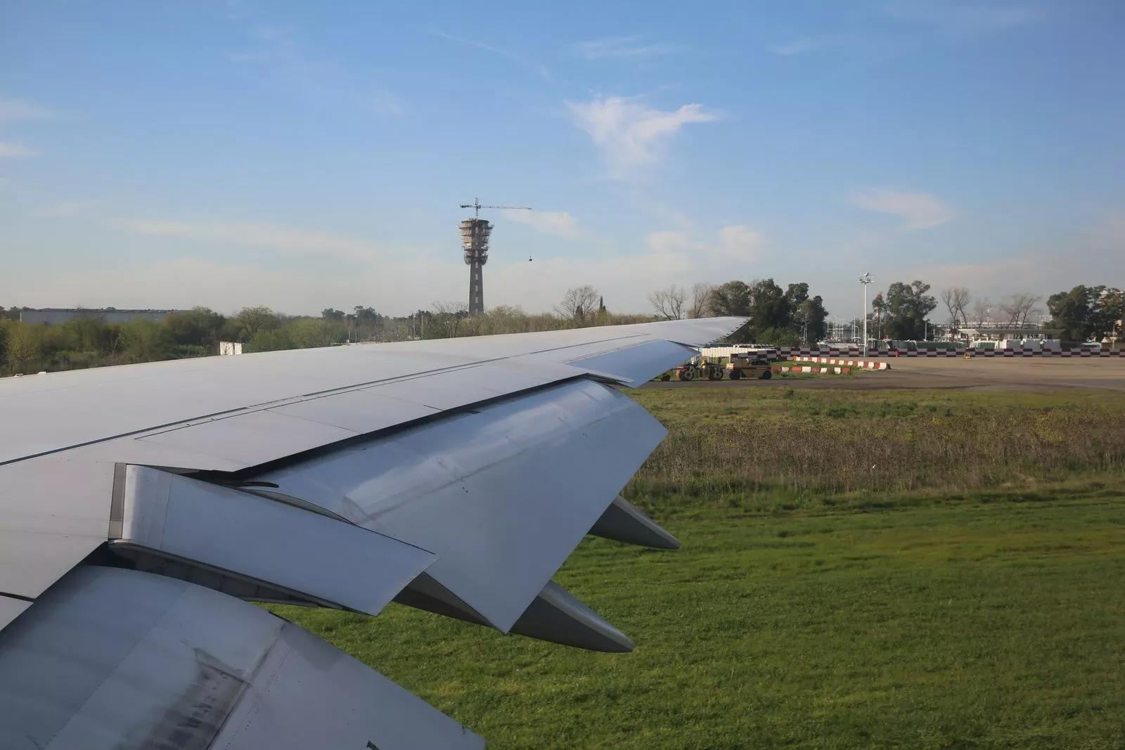 Ala de un avión sobre una calle de rodaje de Ezeiza, con una torre al fondo y pasto a los costados