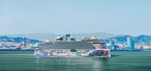 Crucero de casco multicolor navegando frente a la costa de Barcelona, con la ciudad y las montañas al fondo