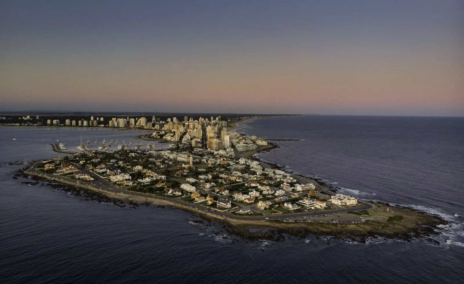 Vista aérea de la península de Punta del Este al atardecer, con edificios junto a la costa y el mar oscuro alrededor