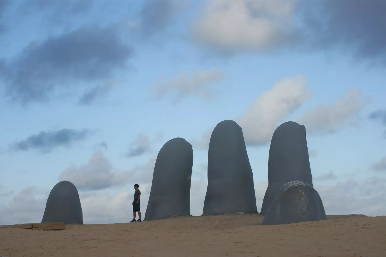 Cuatro dedos de piedra gris que emergen de la arena, con una persona de pie al lado y cielo azul con nubes