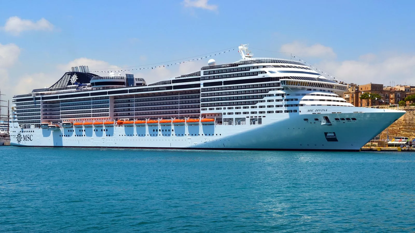 Crucero MSC Divina de casco blanco amarrado en un puerto, con la ciudad de piedra al fondo y cielo azul