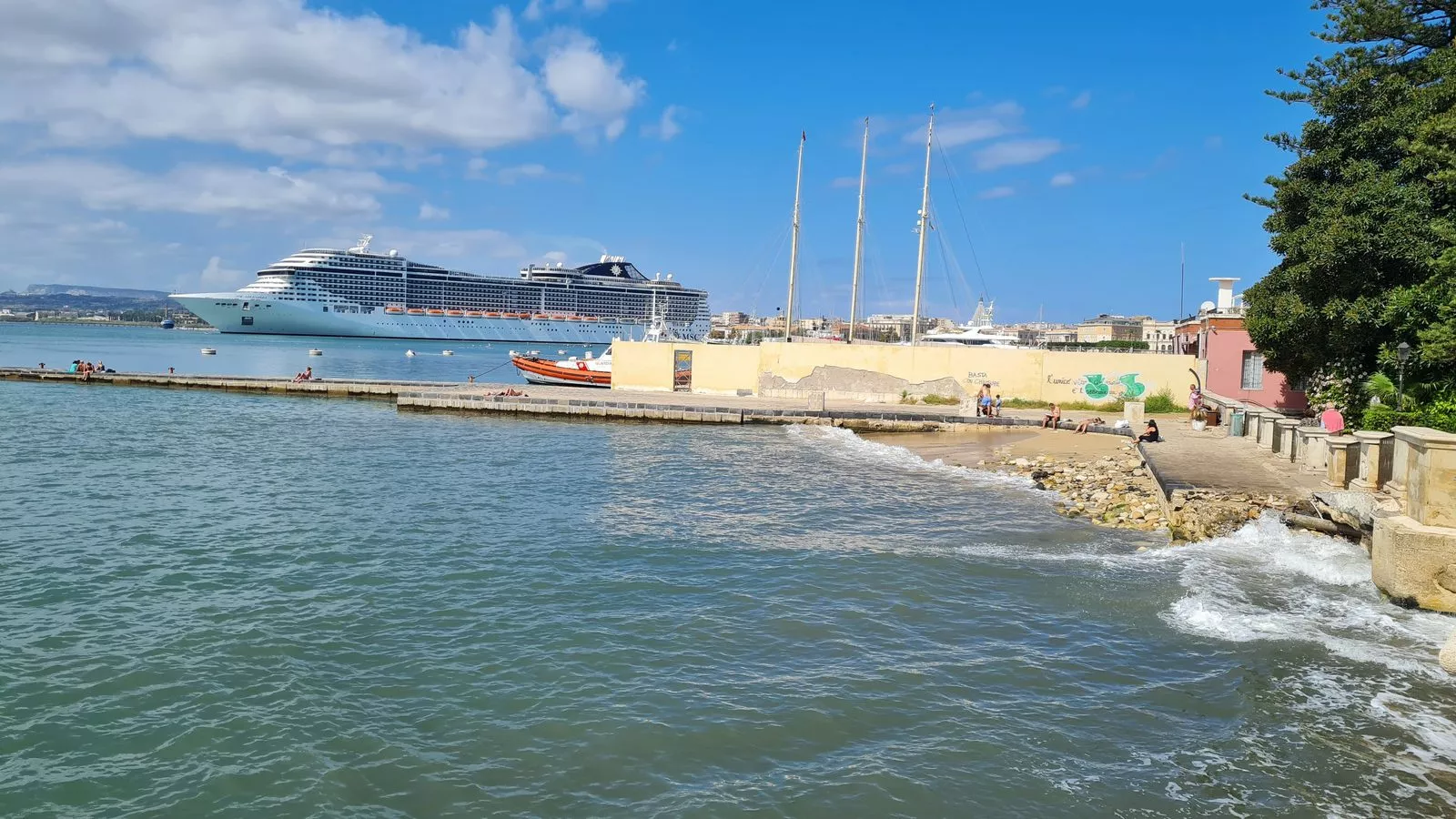 El crucero MSC Splendida amarrado a lo lejos, frente a una costa de rocas y mar de color turquesa